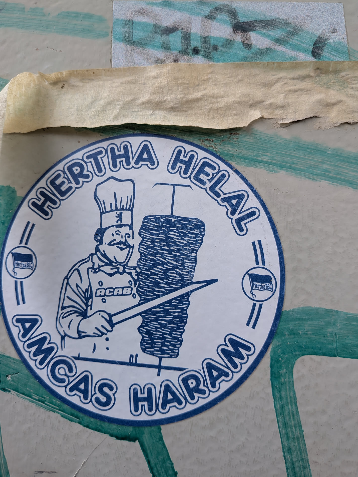 A circular sticker printed in blue ink on a white background. It depicts a cartoon chef with a mustache, wearing a chef's hat adorned with the Berlin bear logo, and an apron with the acronym 'ACAB'. The chef is holding a long knife next to a large spit of doner kebab. The text 'HERTHA HELAL' is arched across the top, and 'AMCAS HARAM' is arched across the bottom. On the left and right sides, there are small circular icons containing the Hertha BSC football club flag.