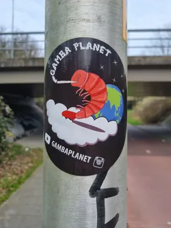 A sticker featuring a cartoon shrimp smoking a cigarette, floating on a cloud in front of the Earth. The text "Gamba Planet" is visible, along with social media icons for YouTube and Instagram.