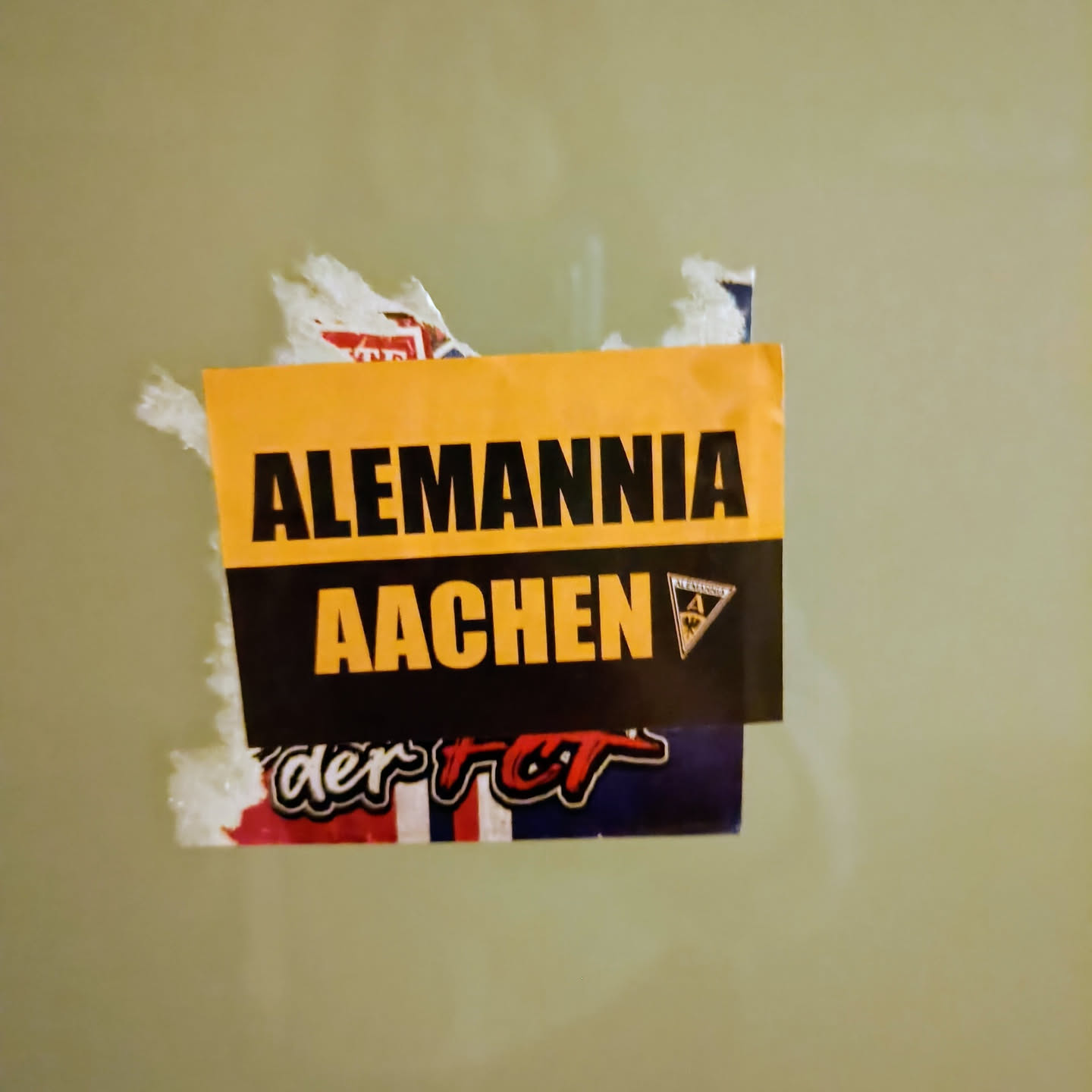 A yellow and black sticker with the text "Alemannia Aachen" on it.  There is a smaller logo visible to the right of the text.  Part of another sticker is visible underneath.