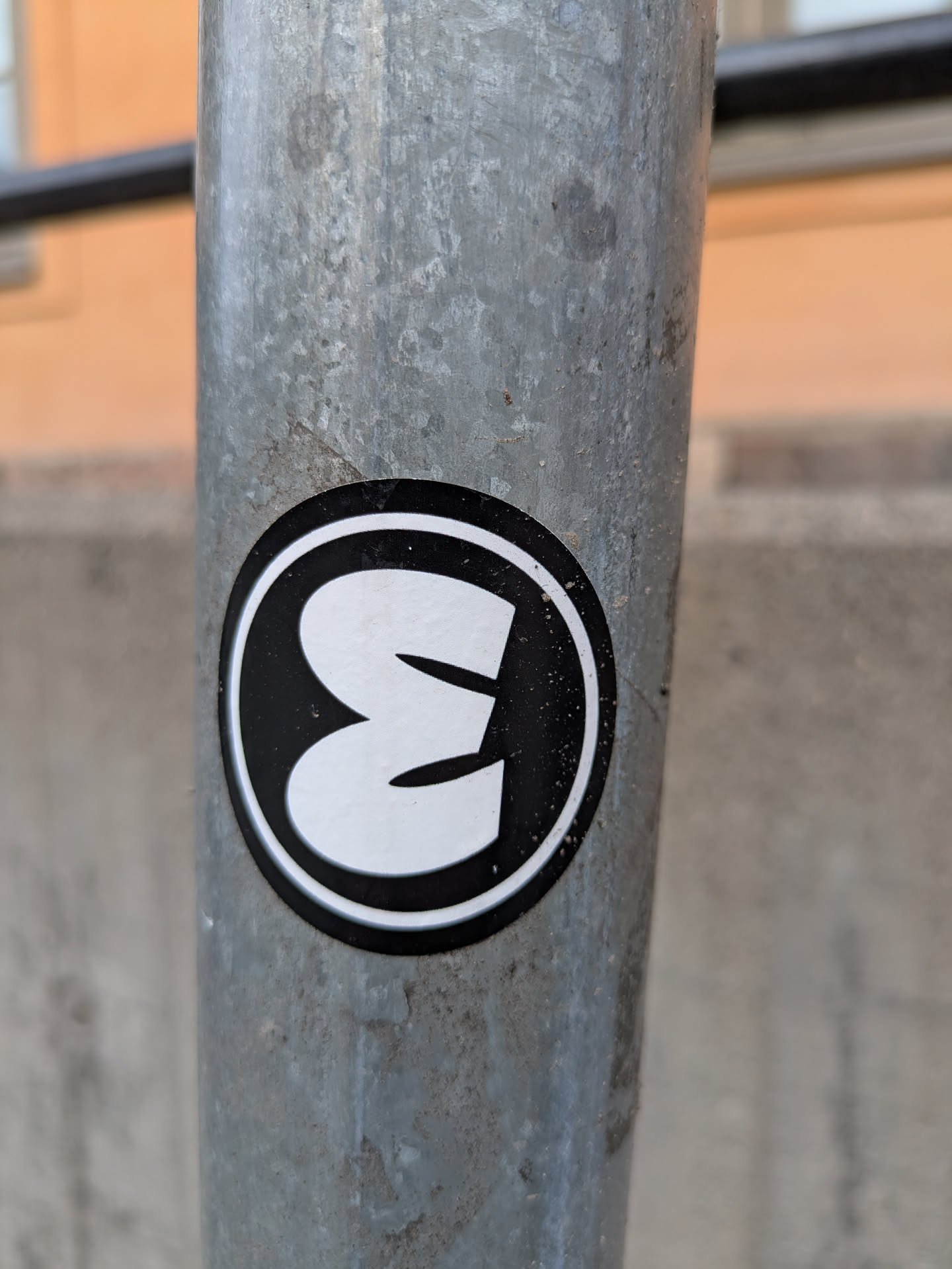 A round black and white sticker affixed to a metal pole. The sticker features a stylized letter 'E' in white against a black background, with a white border around the black circle.
