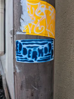 The sticker is rectangular with slightly rounded corners, adhered to a dark, weathered pole. Its design features the word 'DOERKOP' in a bold, dark blue, somewhat blocky font. The letters appear to be spray-painted or stenciled, with a textured, almost pixelated or distressed effect, against a lighter, mottled blue and white background. The overall impression is one of urban graffiti art, with a cool-toned color palette.