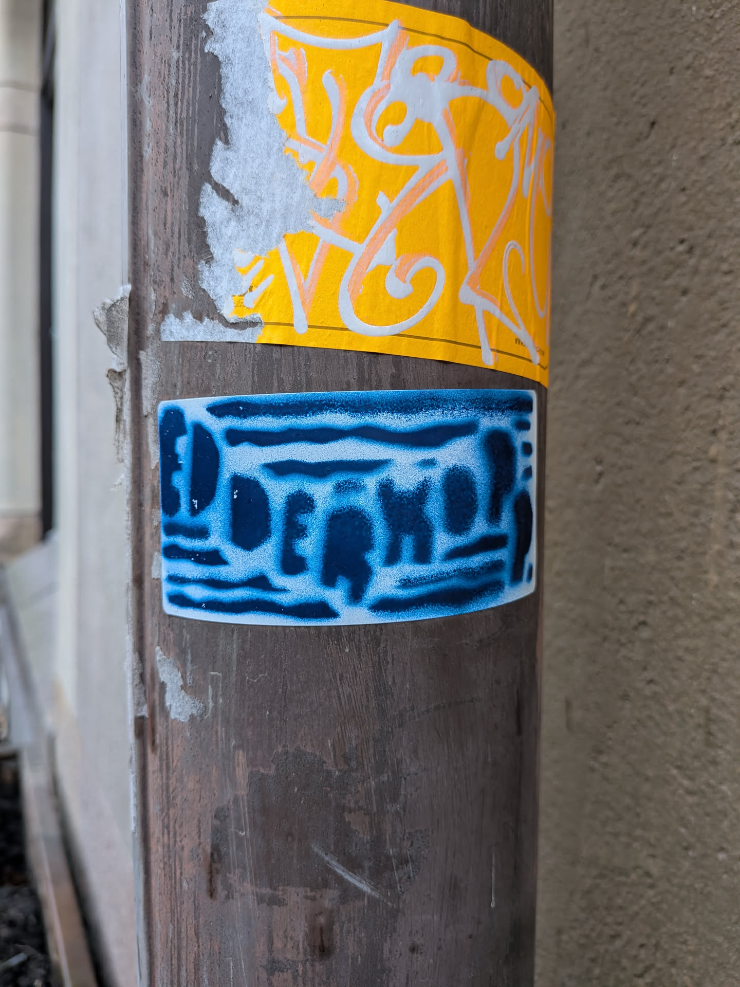 The sticker is rectangular with slightly rounded corners, adhered to a dark, weathered pole. Its design features the word 'DOERKOP' in a bold, dark blue, somewhat blocky font. The letters appear to be spray-painted or stenciled, with a textured, almost pixelated or distressed effect, against a lighter, mottled blue and white background. The overall impression is one of urban graffiti art, with a cool-toned color palette.