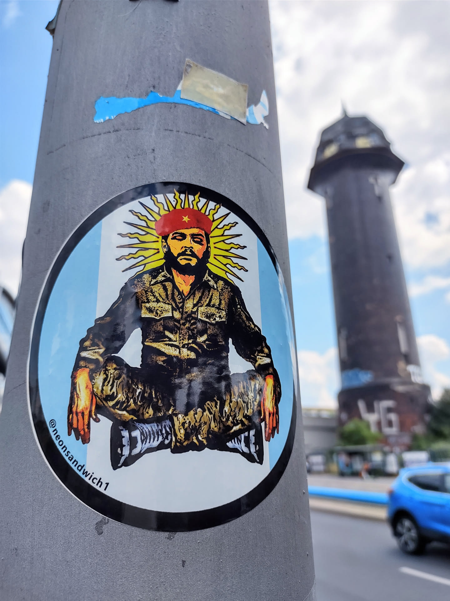 Round sticker featuring a stylized illustration of Che Guevara in a meditative pose, set against a light blue background reminiscent of an Argentinian flag. The artwork is vibrant and bold, with Che depicted wearing his iconic beret and military-style uniform. The sticker is affixed to a grey metal pole, with other smaller stickers visible nearby.