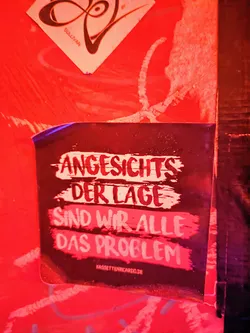 A dark sticker with white text on a red background. The text says "Angesichts der Lage sind wir alle das Problem" which translates to "Considering the situation, we are all the problem". There is also a website address at the bottom: kassettenricardo.de