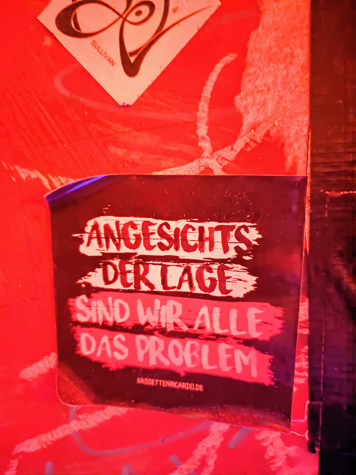 A dark sticker with white text on a red background. The text says "Angesichts der Lage sind wir alle das Problem" which translates to "Considering the situation, we are all the problem". There is also a website address at the bottom: kassettenricardo.de