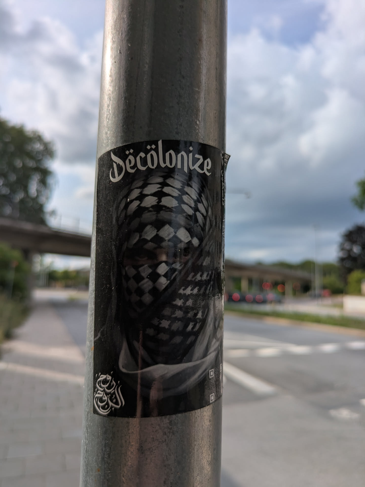 The sticker is rectangular, predominantly black and white/grayscale, and affixed to a metal pole. Its central image is a person's face, mostly obscured by a checkered keffiyeh, with only their eyes visible, conveying a sense of mystery and intensity. Above the figure, in a distinctive gothic-style white font, is the word 'Décolonize'. In the bottom-left corner, there is intricate white Arabic calligraphy. The overall aesthetic is stark and impactful, utilizing high contrast.