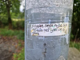 A rectangular sticker affixed to a weathered metal pole. The sticker features hand-written text in Swedish which reads: "Ju djupare sorgen mer slatt sig i dig desto mer tycka kan du rymma."  which roughly translates to:  "The deeper the sorrow, the more you can embrace it." A small sun-like drawing is in the bottom right corner.