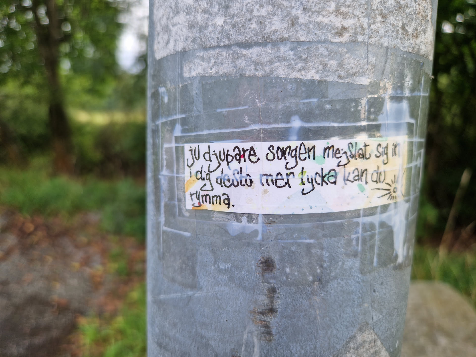 A rectangular sticker affixed to a weathered metal pole. The sticker features hand-written text in Swedish which reads: "Ju djupare sorgen mer slatt sig i dig desto mer tycka kan du rymma."  which roughly translates to:  "The deeper the sorrow, the more you can embrace it." A small sun-like drawing is in the bottom right corner.