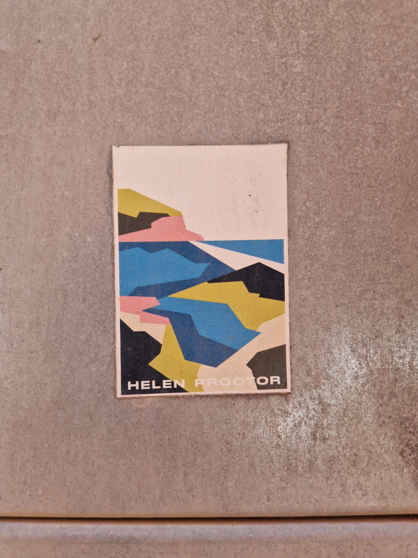 A minimalist sticker featuring an abstract geometric interpretation of a coastal scene. The design is primarily composed of blocks of blue, yellow, pink, and black, suggesting water, land, and sky.  The artist's name, "Helen Proctor," is printed at the bottom.