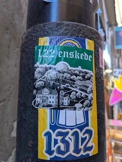A rectangular sticker adhered to a metal post, placed on top of another sticker. The upper section contains a green rectangular banner with stylized white lettering reading '122 enskede'. The lower part contains a detailed black-and-white graphic illustration of Enskede in Stockholm, featuring the historic Enskede gård manor house, lush trees, a metro train line with a sign reading 'TUNNELBANA NR 19 STATION ENSKEDE GÅRD', and the spherical silhouette of the Globe Arena (Globen) along the skyline.
