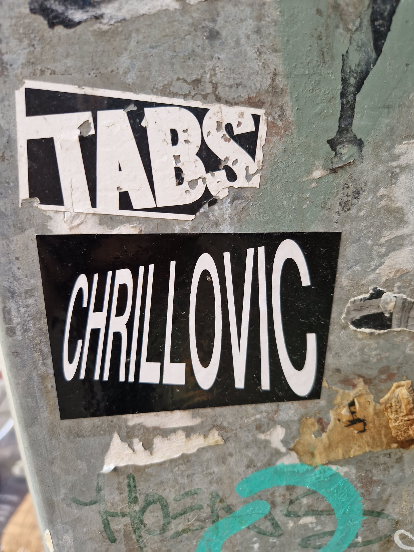 A black sticker with the word CHILLOVIC written in white uppercase letters.  There is a smaller, damaged sticker above it that says TABS. Both are affixed to a weathered surface.