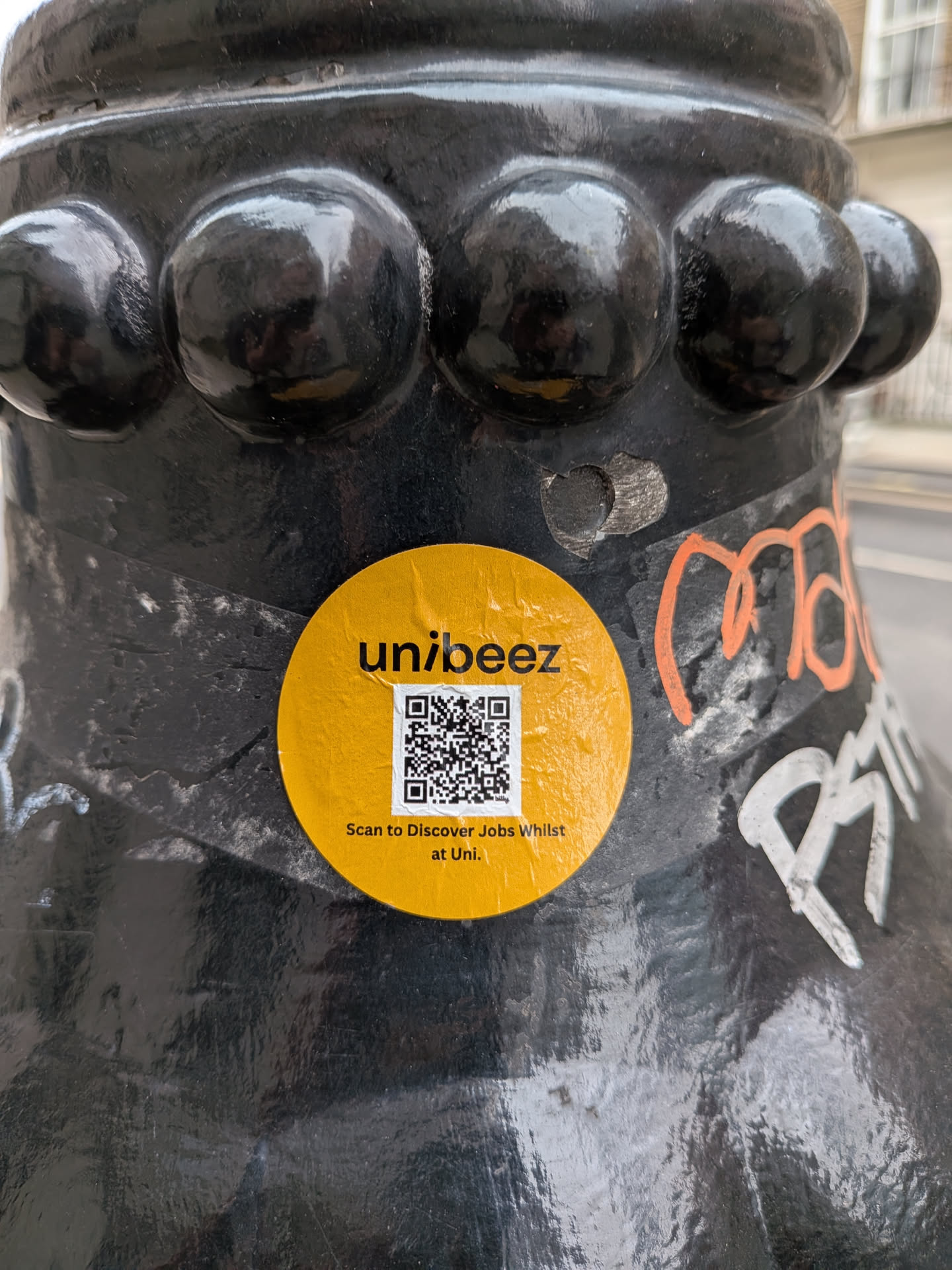A circular, bright mustard yellow sticker featuring the brand name 'unibeez' in black, lowercase sans-serif font at the top. Below the brand name, a prominent black and white QR code is centered. Underneath the QR code, in smaller black text, it reads 'Scan to Discover Jobs Whilst at Uni.' The sticker is applied to a dark, textured, possibly black metal surface, which appears to be a bollard or lamppost, showing signs of wear and other graffiti around it.
