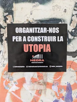 The sticker, adhered to a weathered wall, features bold white text against a black background, with the word 'UTOPIA' highlighted in coral. Below the text is the logo for Hedra, an anarchist organization, followed by contact information. The wall around the sticker is covered in graffiti and peeling paint, adding to the sticker's rough, urban aesthetic.
