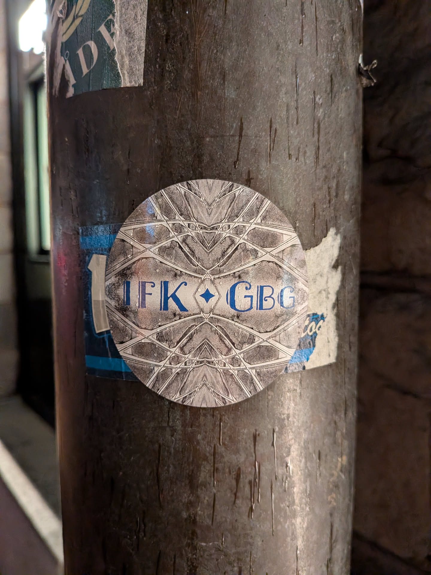 The sticker is circular and features a highly symmetrical, intricate geometric pattern in shades of gray and white. The pattern resembles a stylized network of lines and diamonds. In the center, the text 'IFK GBG' is prominently displayed in a bold, blue, sans-serif font. A small blue diamond shape is positioned between 'IFK' and 'GBG'. The sticker appears to be adhered to a rough, textured surface, possibly a metal pole, with some surrounding peeling paper.