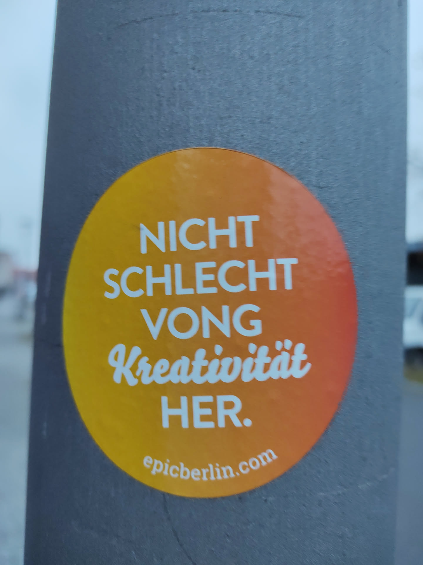 Round orange sticker with the text "Nicht schlecht von Kreativität her." and the website epicberlin.com at the bottom. The sticker is on a gray pole.