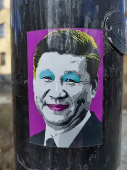 A sticker featuring a pop art rendition of Xi Jinping, the president of China.  The image is stylized with bold colors and makeup, creating a striking contrast to the usual formal depictions. The sticker is affixed to a dark pole.