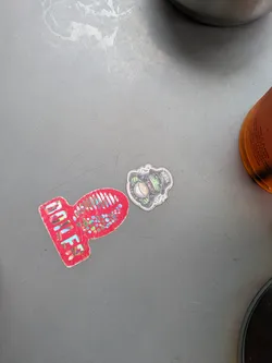 This small sticker has a dark green border and a grey and green image inside. The image features a large, detailed eye with what appears to be plant life growing around it, giving it a somewhat unsettling and organic feel.
