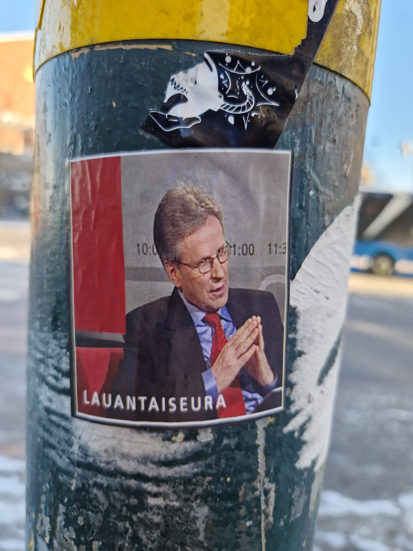 A sticker featuring a photo of a man in a suit, with the word "LAUANTAISEURA" at the bottom.  The sticker is affixed to a pole.