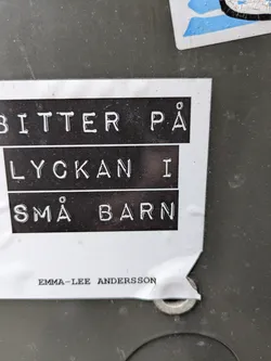 A sticker with the text "Bitter på lyckan i små barn" in Swedish, which translates to "Bitter on happiness in small children."  The sticker also includes the artist's name: Emma-Lee Andersson.