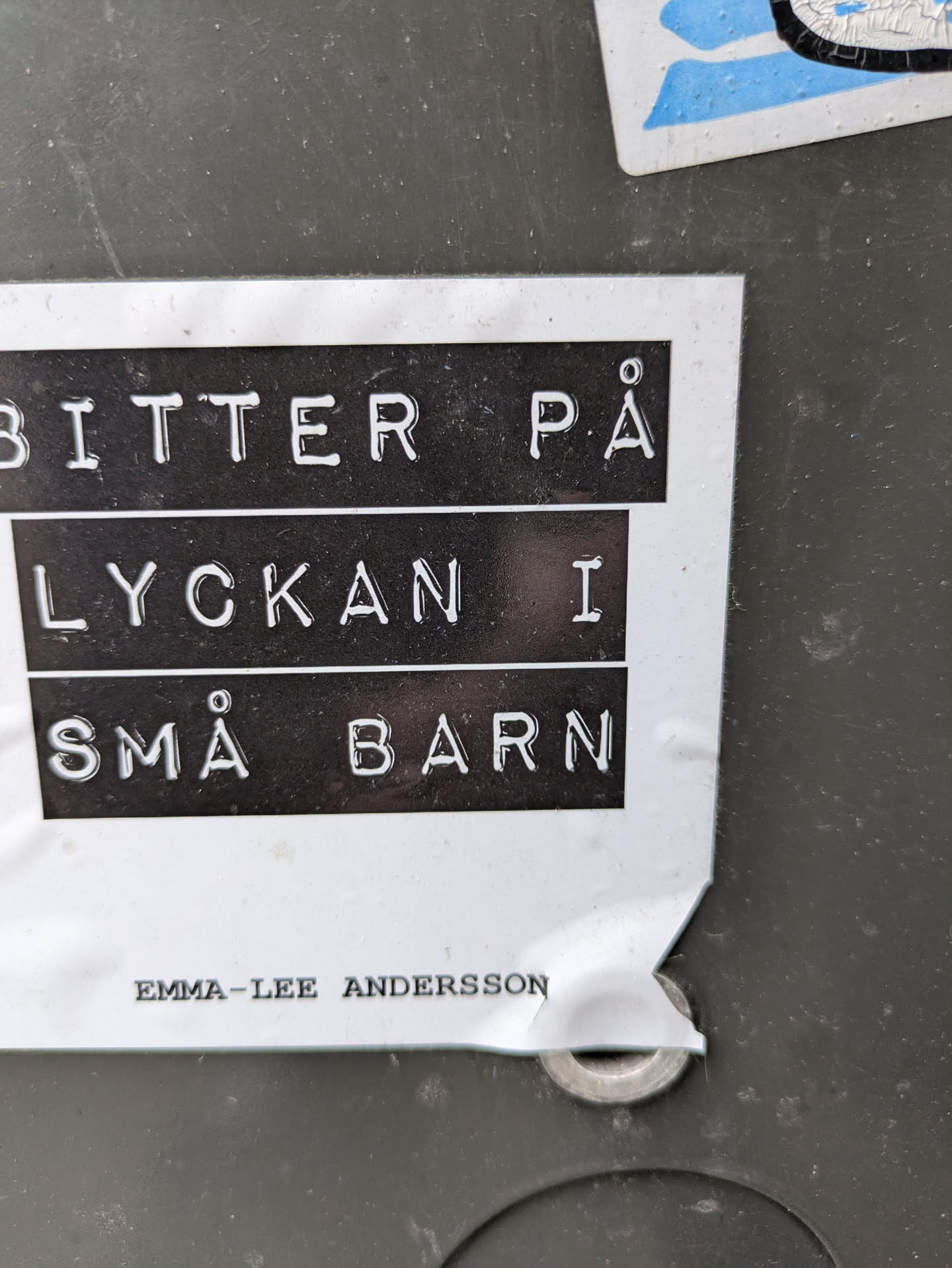 A sticker with the text "Bitter på lyckan i små barn" in Swedish, which translates to "Bitter on happiness in small children."  The sticker also includes the artist's name: Emma-Lee Andersson.