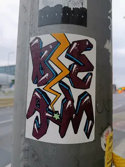 A vibrant sticker featuring stylized lettering, 'KSAM', with a lightning bolt graphic. The color palette is a combination of maroon, yellow, and teal. The style is reminiscent of graffiti or street art.