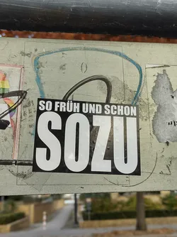 A black and white sticker with the text "SO FRÜH UND SCHON SOZU" in bold white letters on a black background. The sticker is affixed to a surface with other stickers and graffiti. The background is blurred, showing a street scene.
