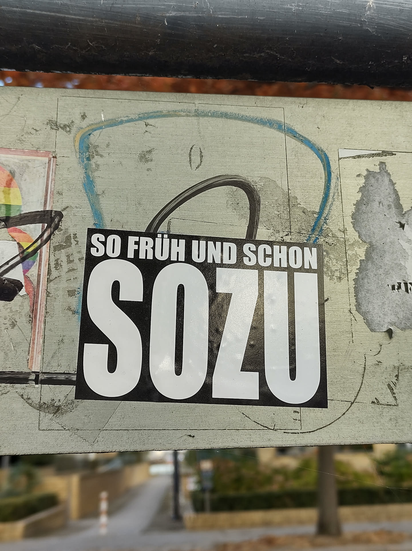 A black and white sticker with the text "SO FRÜH UND SCHON SOZU" in bold white letters on a black background. The sticker is affixed to a surface with other stickers and graffiti. The background is blurred, showing a street scene.