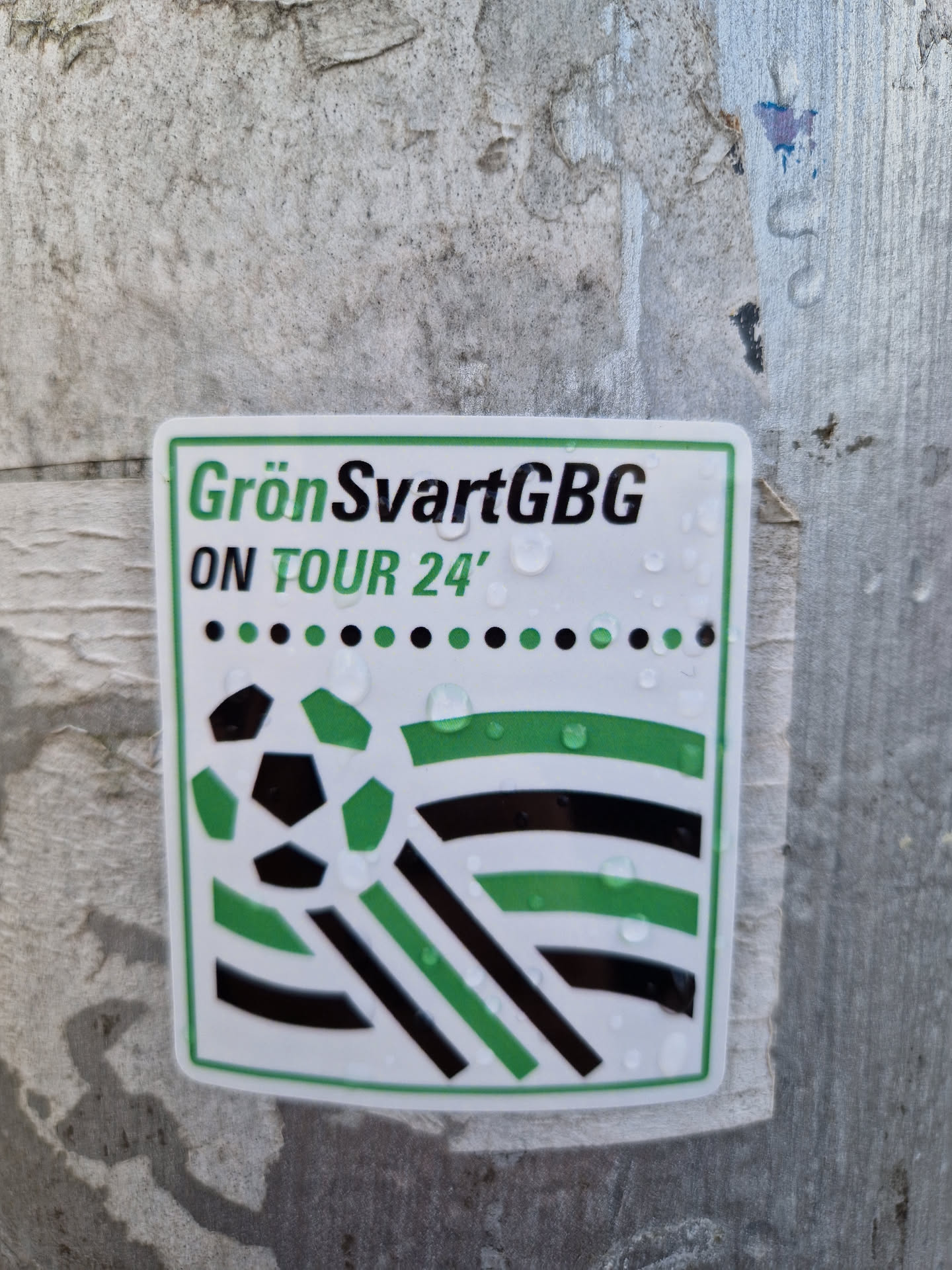 A sticker featuring the text 'GrönSvartGBG ON TOUR 24' and a graphic design with a soccer ball and green and black stripes. The sticker appears to be affixed to a weathered surface, with water droplets visible on its surface.