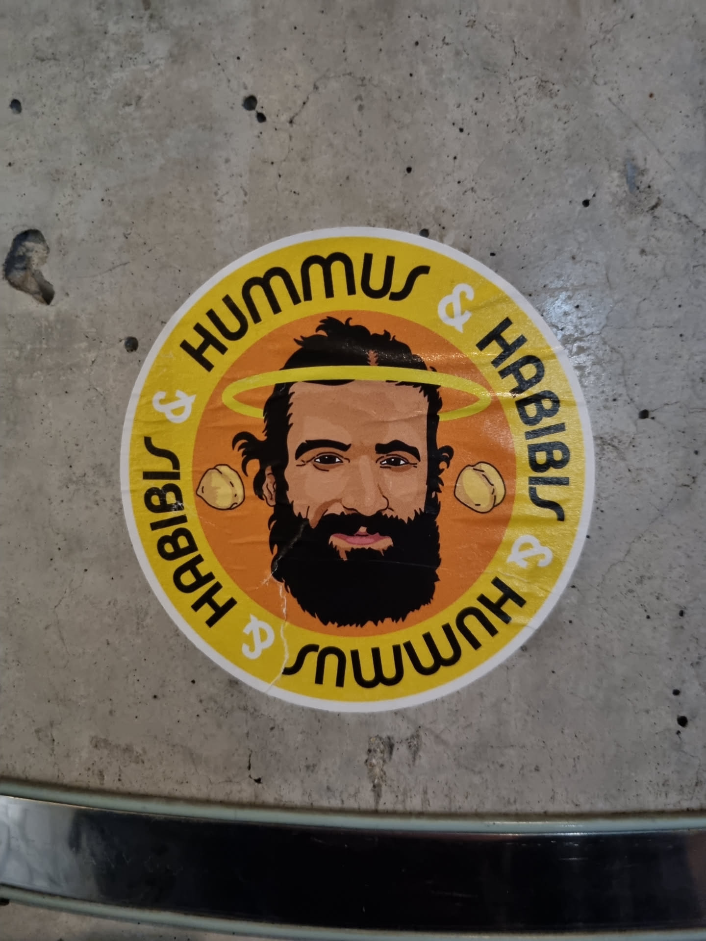 Round sticker featuring a cartoon illustration of a man with a beard and halo, surrounded by text that reads Hummus & Habib's. The background is orange, and the text is in yellow.