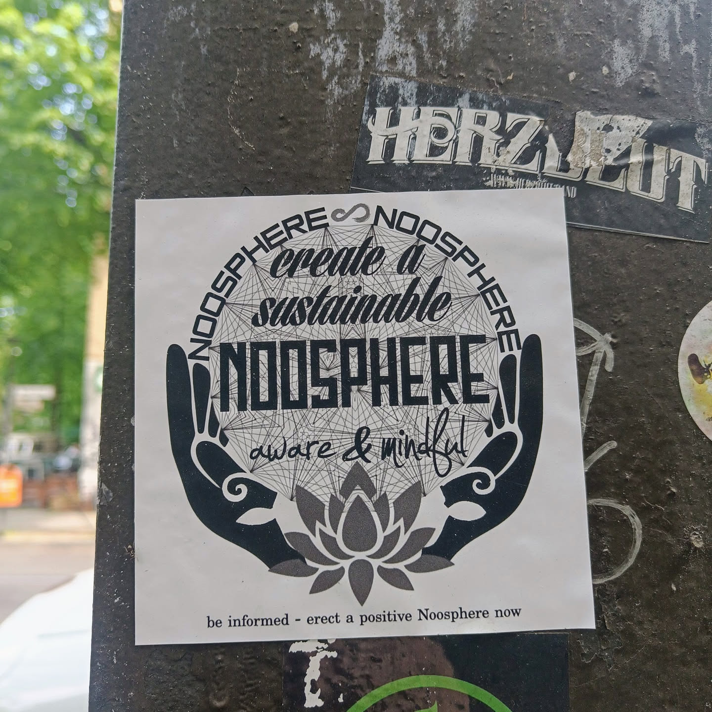A sticker with a circular design featuring two hands holding a lotus flower inside a web-like pattern. The words "NOOSPHERE create a sustainable NOOSPHERE aware & mindful" are written around the design. At the bottom, it says: "be informed - erect a positive Noosphere now".