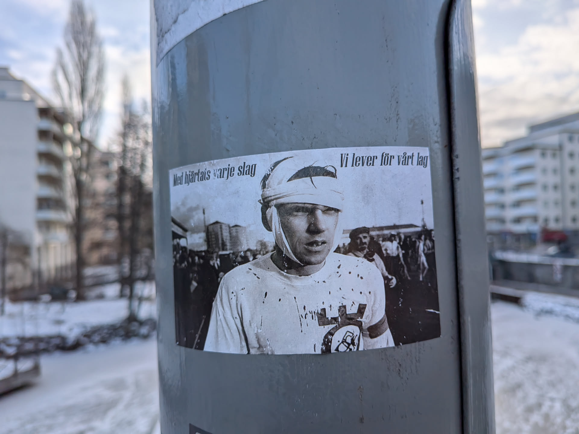 A rectangular, horizontally oriented sticker featuring a black and white photograph. The central figure is a young man, likely a football player, with a bandaged head covering his eyes and forehead. He wears a light-colored t-shirt splattered with dark marks, possibly blood or mud. On his right arm and chest, a circular emblem with a stylized 'AIK' logo is visible. In the blurred background, a crowd of spectators and some industrial-looking structures can be seen. The overall aesthetic is gritty and evokes a sense of historical struggle or intense dedication.