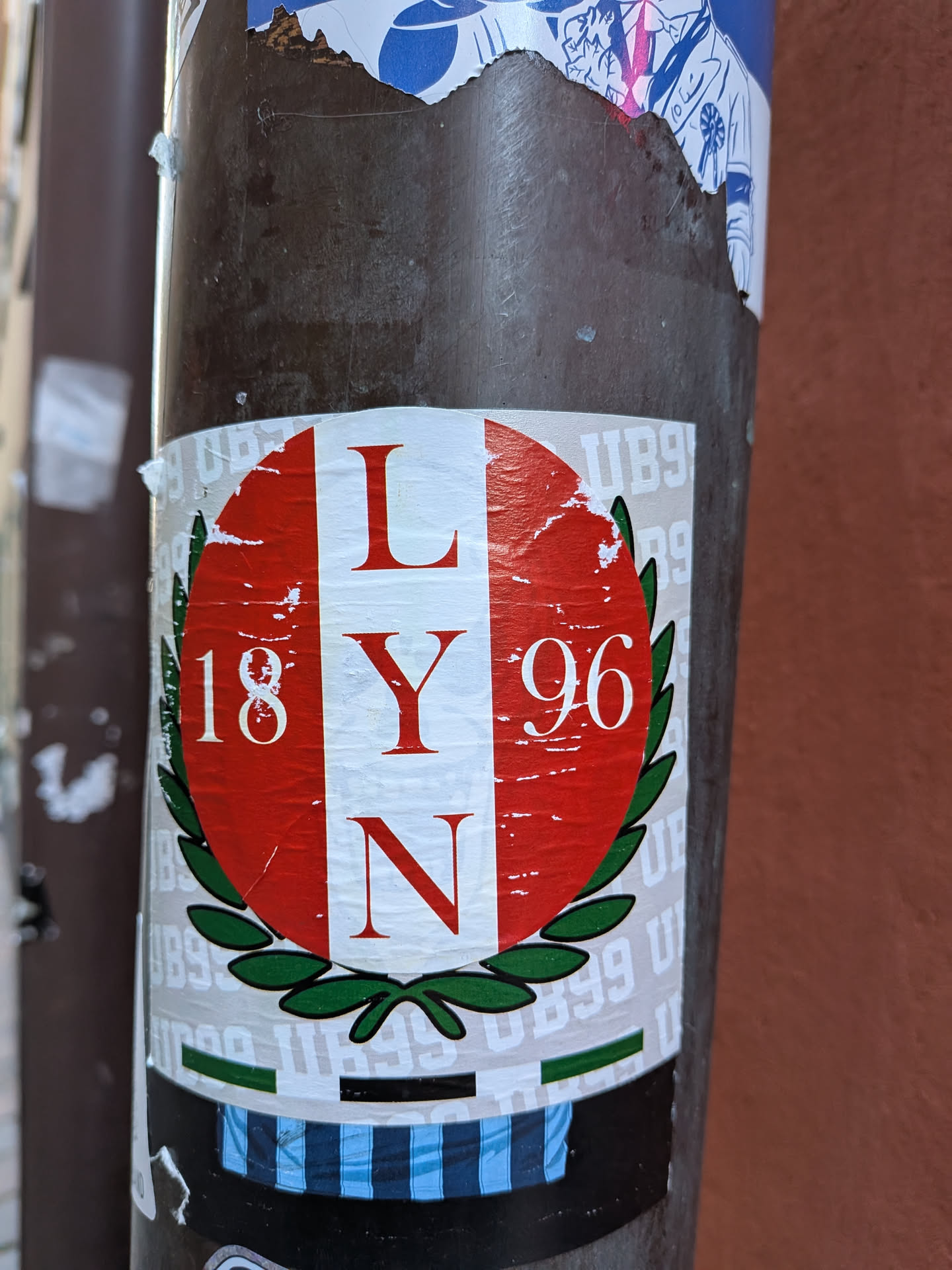 The central sticker is circular, featuring a vertical red-white-red stripe pattern, reminiscent of a flag. Centered on the white stripe are the red capital letters 'L', 'Y', and 'N' stacked vertically in a serif font. To the left, on the red background, are the white numbers '18', and to the right, '96', also in a serif font. The lower half of the circular design is framed by a green laurel wreath. The sticker is applied to a dark brown pole and shows signs of wear, including creases and minor tears. Below the main circular design, a horizontal band with blue and white vertical stripes is visible. The main sticker is set against a larger, rectangular white sticker, which itself covers other stickers, one of which appears to have 'UB99' repeated in its pattern.