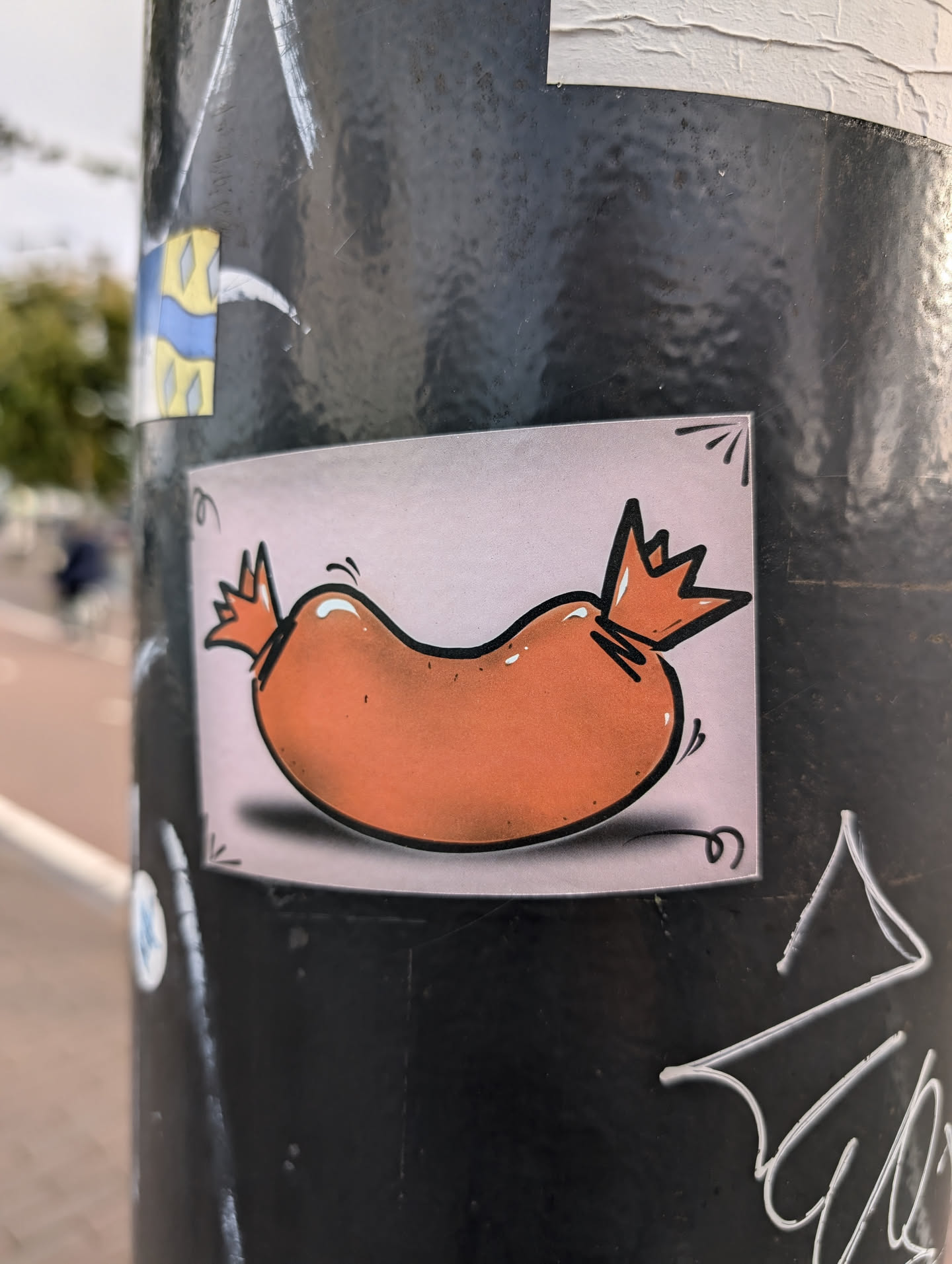 A sticker of a cartoon sausage with a crown on each end. The sausage is orange and the background is light pink. The sticker is placed on a dark gray pole.