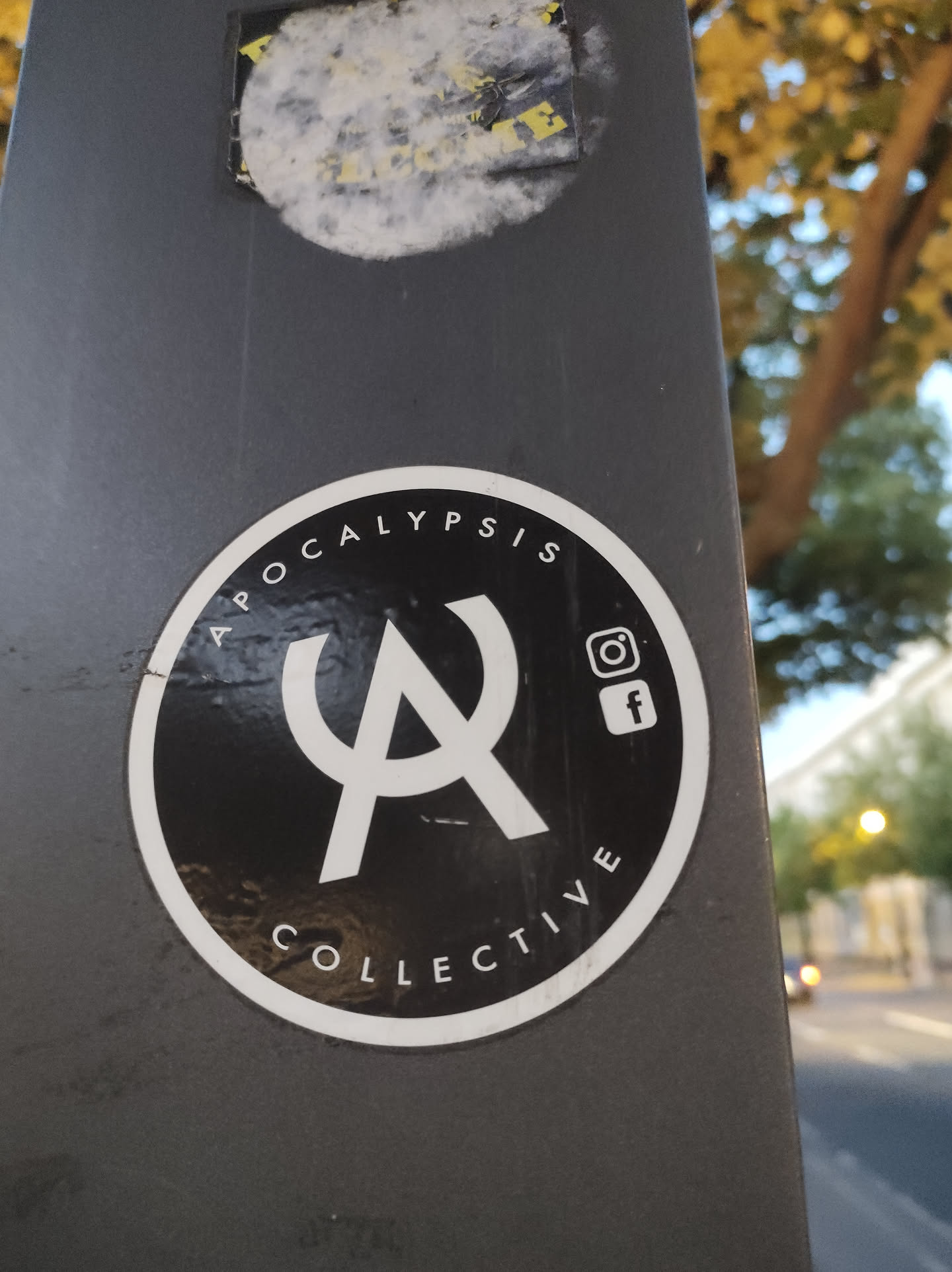 A round black and white sticker with the text 'Apocalypse Collective' and a stylized 'A' logo.  Instagram and Facebook icons are also present. The sticker is affixed to a dark grey pole.