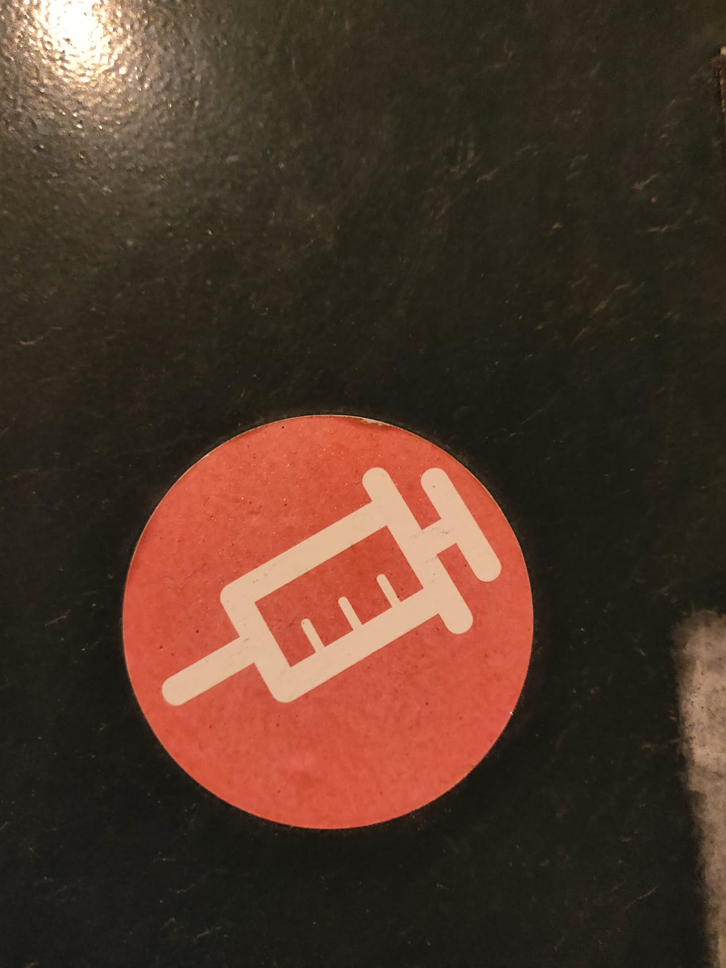 Round, coral-colored sticker featuring a white outline of a syringe.  The design is minimalist and the color contrast is striking against most backgrounds.