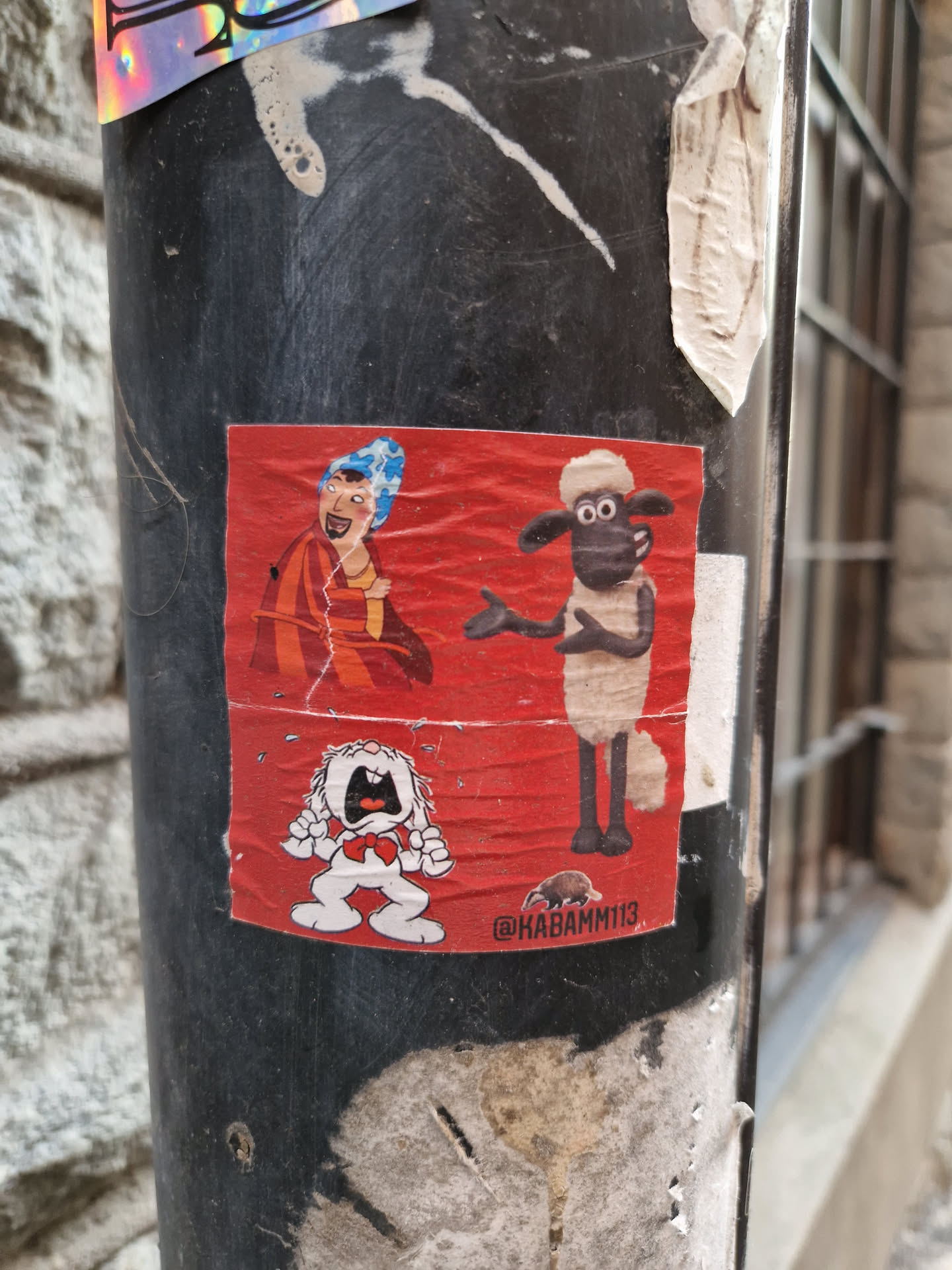 A red sticker featuring Shaun the Sheep, a woman with blue hair, and a crying dog.  The sticker has a small illustration of an anteater and an Instagram handle at the bottom. It is affixed to a dark-colored pole.