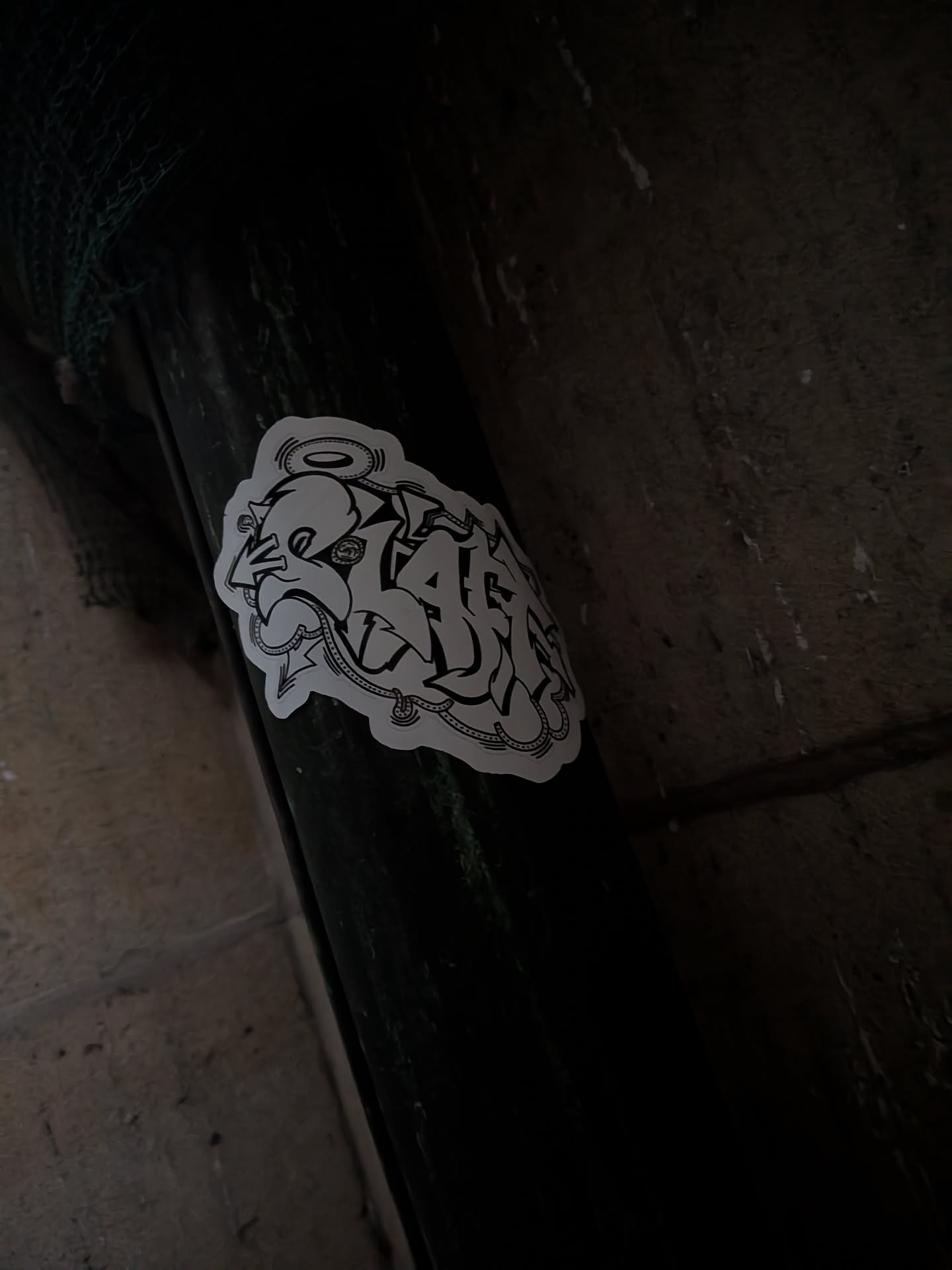 A die-cut sticker placed on a dark post or pipe. The sticker features bold black line art on a white background forming stylized graffiti letters, possibly spelling 'SLAFF'. It is decorated with an arrow on the left, a halo floating above the first letter, and a dotted chain-like embellishment curving along the bottom edge.
