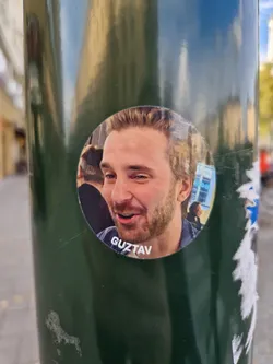 Round sticker featuring a portrait of a man named Guztav. The sticker is affixed to a dark green pole outdoors.