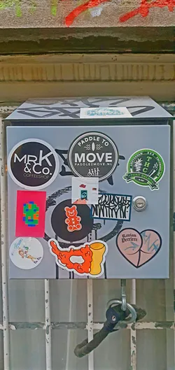 This is a metal box covered in an assortment of stickers. The stickers have various designs and logos, showcasing different brands, art styles, and themes. The box itself is grey, and the stickers add pops of color and visual interest to the otherwise plain surface.