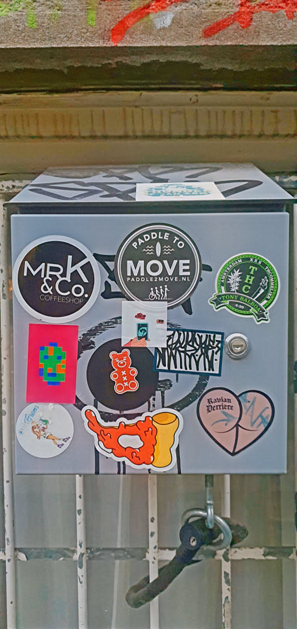 This is a metal box covered in an assortment of stickers. The stickers have various designs and logos, showcasing different brands, art styles, and themes. The box itself is grey, and the stickers add pops of color and visual interest to the otherwise plain surface.