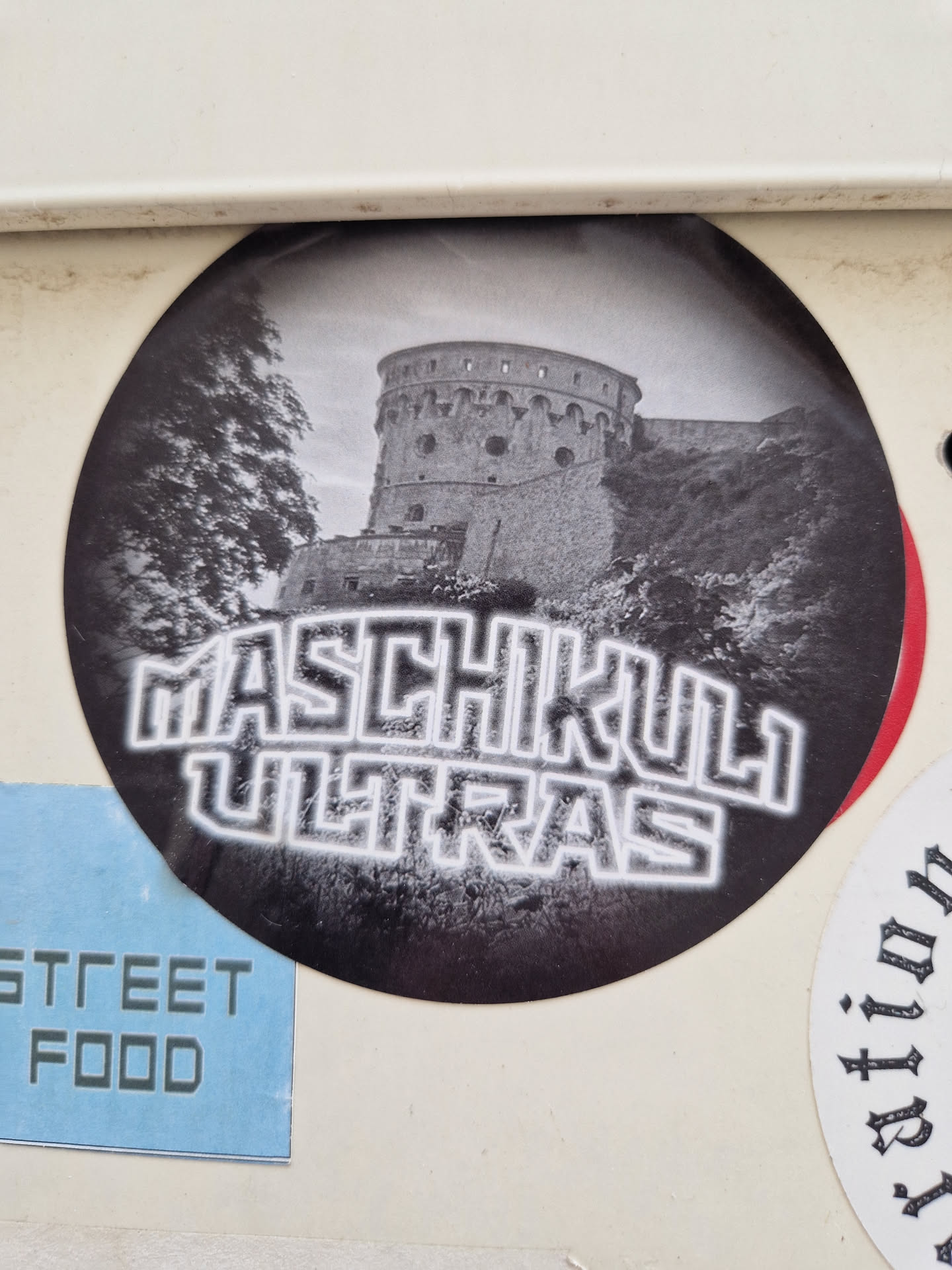 Round sticker with a black and white illustration of a castle, with the text 'Maschikuli Ultras' in a stylized font below it. The style is reminiscent of a band logo or urban art. 