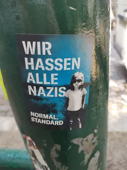 A sticker with a girl wearing sunglasses on a blue background. The text 'Wir hassen alle Nazis' (We hate all Nazis) is written in bold white letters. At the bottom, it says 'NORMAL STANDARD'.