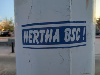 This is a rectangular sticker with rounded corners featuring a blue background and white text that reads "HERTHA BSC!" It's placed on a white pole. There are two white lines on the top and bottom of the text box.