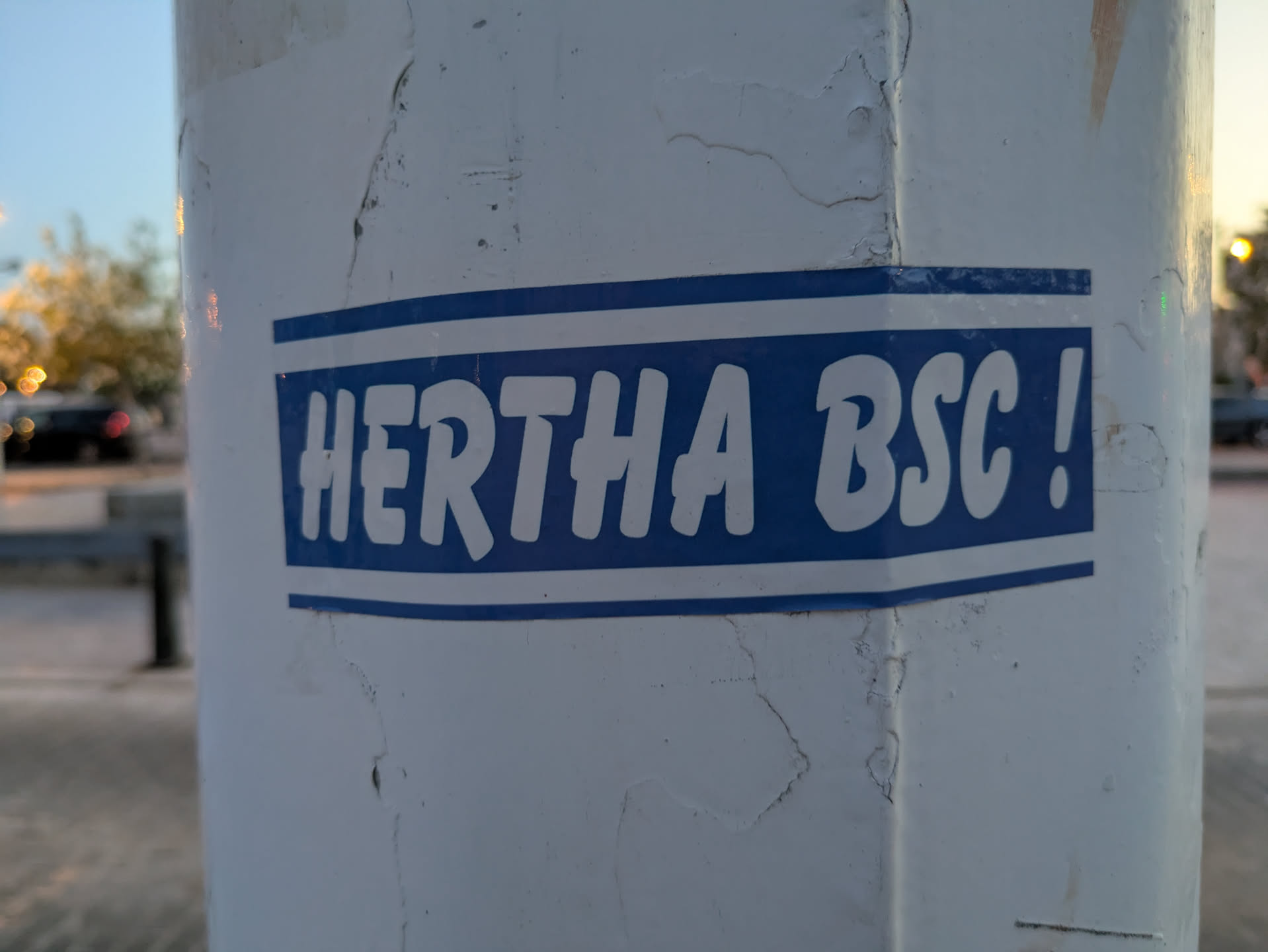 This is a rectangular sticker with rounded corners featuring a blue background and white text that reads "HERTHA BSC!" It's placed on a white pole. There are two white lines on the top and bottom of the text box.