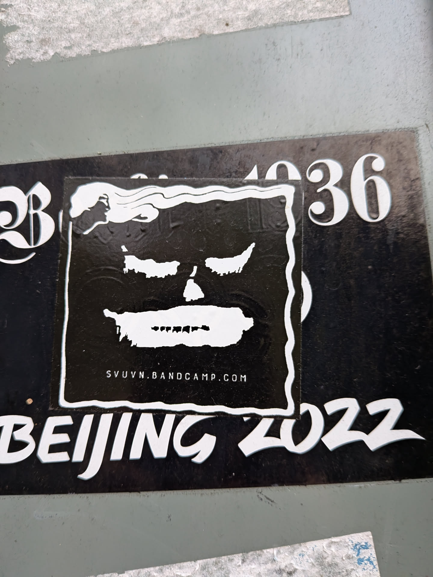 A square, high-contrast black sticker featuring a stylized, shadowy face in white. The face has squinted eyes, a small nose cavity, and a smeared white mouth with jagged hints of teeth. A wavy white border frames the design, curving at the top left into fluid, cloud-like ripples. Below the face, the URL 'SVUVN.BANDCAMP.COM' is printed in small, clean uppercase lettering.