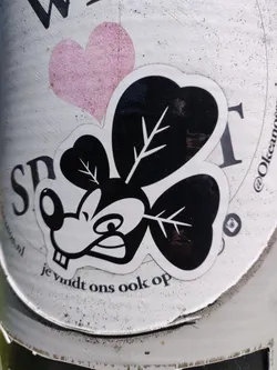 A black and white sticker featuring a stylized mouse character with clover-shaped ears. The character has a somewhat grumpy expression.  The sticker is placed on a white surface, partially obscuring other text and a pink heart shape.