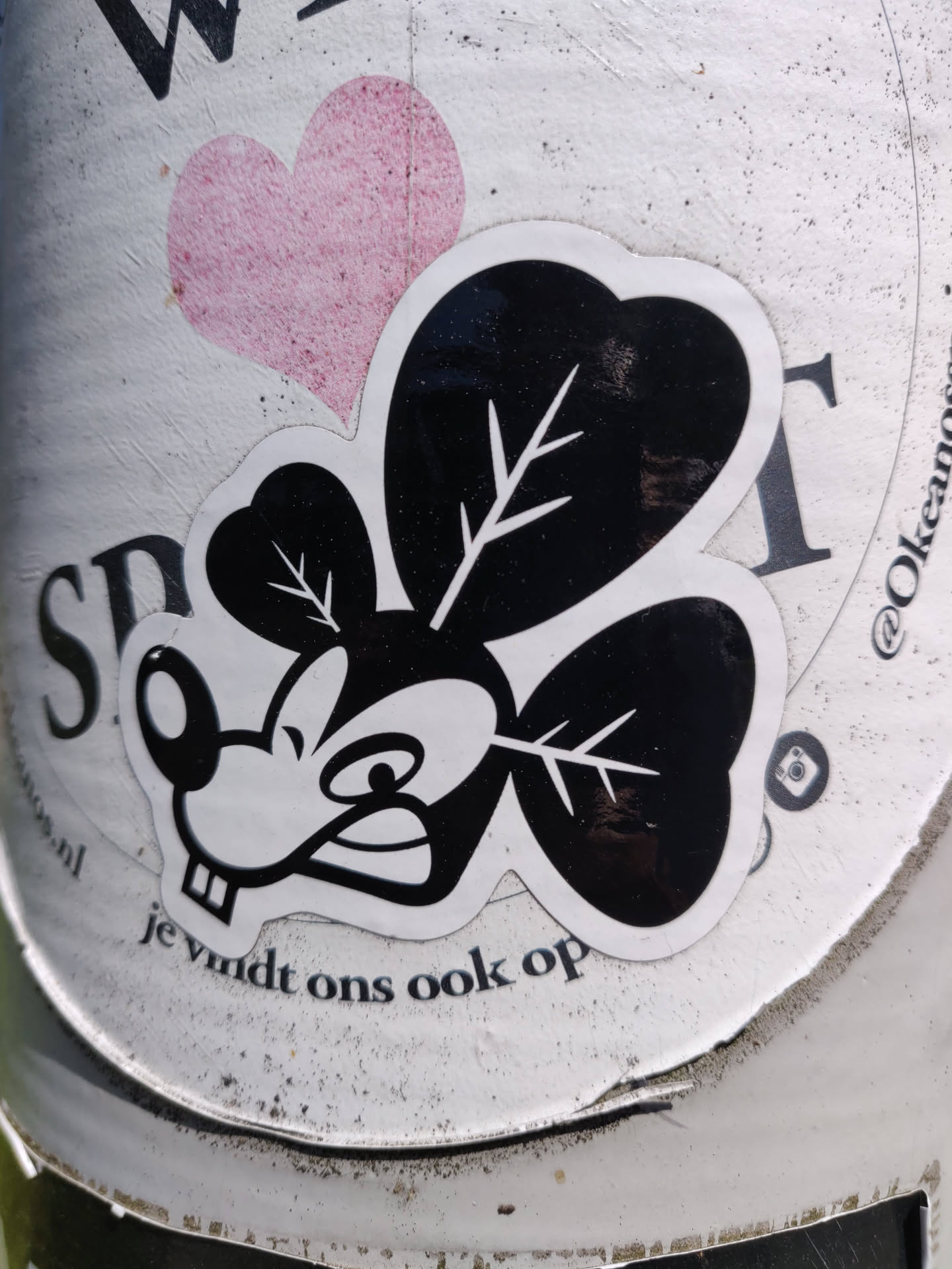 A black and white sticker featuring a stylized mouse character with clover-shaped ears. The character has a somewhat grumpy expression.  The sticker is placed on a white surface, partially obscuring other text and a pink heart shape.