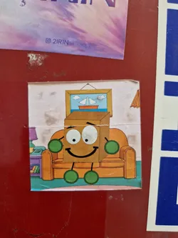 A cartoon sticker featuring a happy box character relaxing on an orange couch in a living room setting. The box has a smiley face and small green limbs. There's a picture of a sailboat on the wall above the couch.