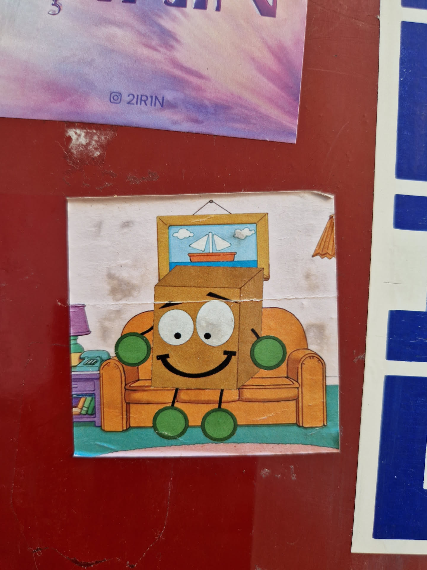 A cartoon sticker featuring a happy box character relaxing on an orange couch in a living room setting. The box has a smiley face and small green limbs. There's a picture of a sailboat on the wall above the couch.