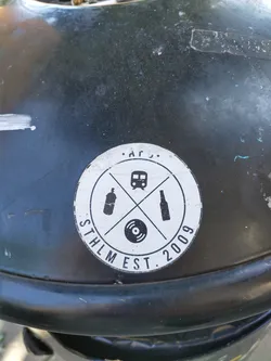 A round black and white sticker with a logo. The logo features a train, a spray can, a bottle and a vinyl record crossed in an 'x' shape. The text 'STHLM EST. 2009' is visible.  It appears to be a logo for a Stockholm-based establishment founded in 2009, perhaps a bar, club or record store.